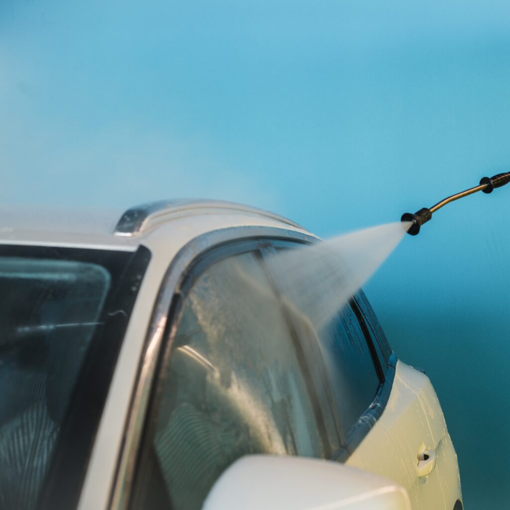 Unrecognizable female client spraying water on windows of modern vehicle with jet hose against blue wall in self service car wash facility